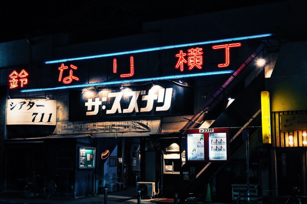 Eine stimmungsvolle japanische Kinofassade bei Nacht mit leuchtenden Neonbuchstaben und Filmplakaten.