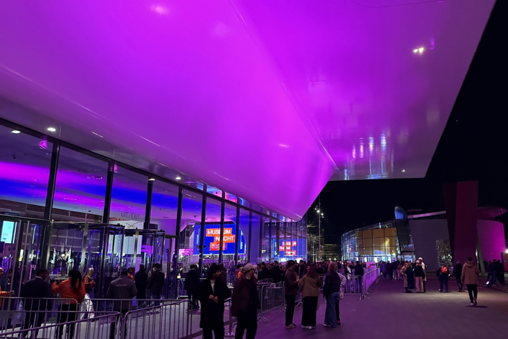 Der Eingang zum Stedelijk Museum Amsterdam bei Nacht, beleuchtet von violetten Lichtern, mit Menschen, die für die Museumsnacht in der Schlange stehen.
