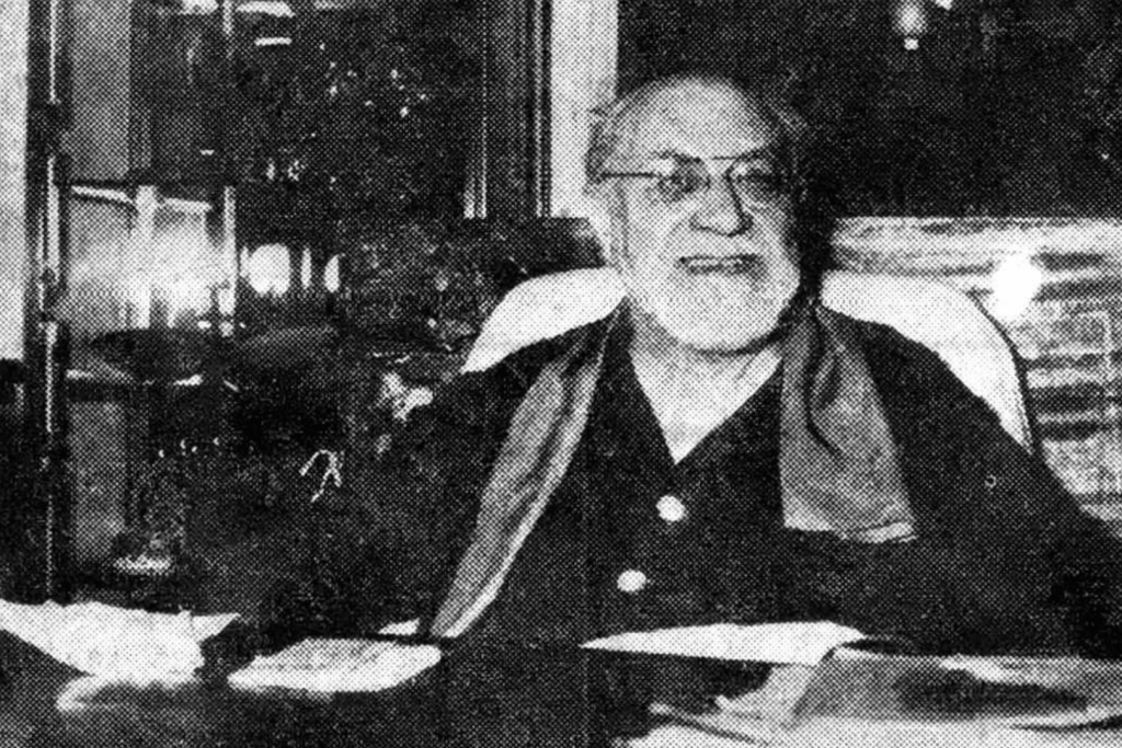 Schwarz-Weiß-Archivfoto eines älteren Henri Matisse mit Brille und Bart, der an seinem Arbeitstisch sitzt.
