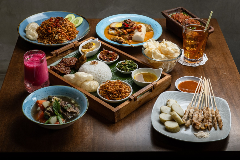 Ein Holztisch, reich gedeckt mit einem traditionellen indonesischen Reisgericht, frischem Hähnchen-Saté, Lontong, Bami Goreng und erfrischenden Getränken