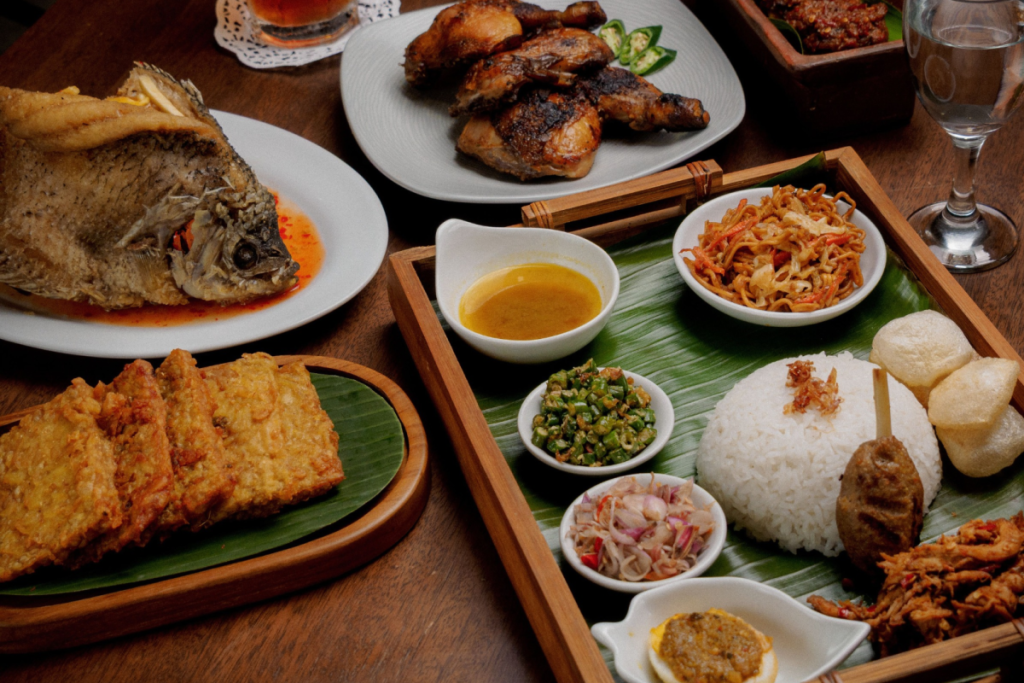 Authentische indonesische Gerichte, darunter knusprig gebratener Fisch, Tempeh, Gado-Gado, Nasi Putih und Rendang, serviert auf einer Holzschale.