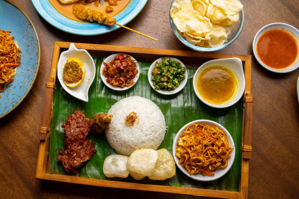 Draufsicht auf ein reichhaltiges indonesisches Reisgericht auf einem Holztablett mit Bananenblatt, frischem Sambal, Mie Goreng und Rendang.