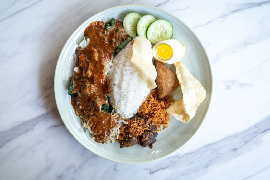 Ein Teller mit einem authentischen indonesischen Gericht, bestehend aus weißem Reis, Gado-Gado mit Erdnusssoße, zartem Rendang, Gurke, gebratenen Zwiebeln und einem halben Ei.