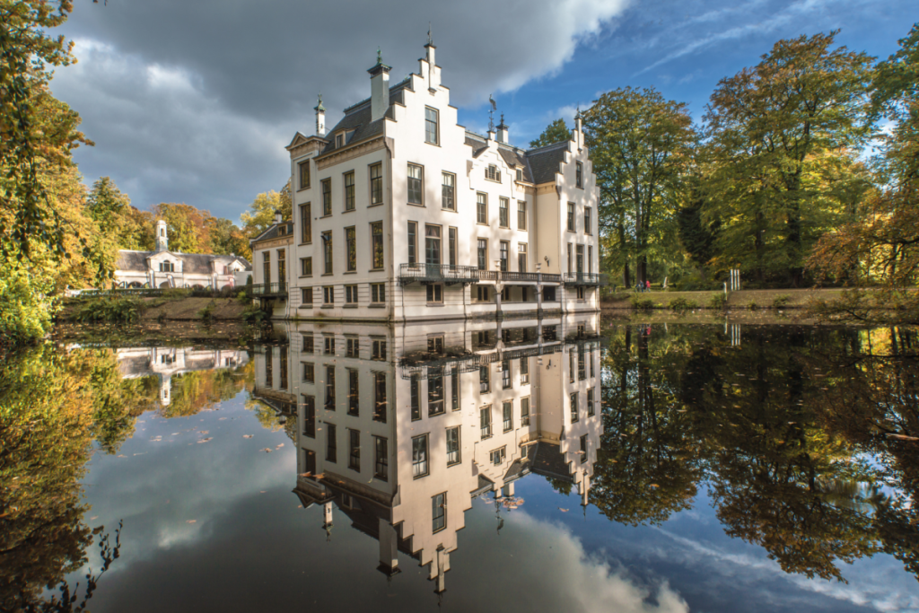 Das weiße Schloss Staverden spiegelt sich im stillen Wasser des Schlossgrabens wider, umgeben von Herbstbäumen auf der Veluwe.