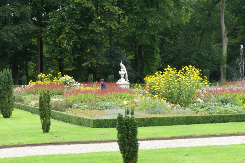 Farbenfroher Blumengarten auf dem Landgut Staverden mit der ikonischen Statue eines weißen Pfaus im Hintergrund