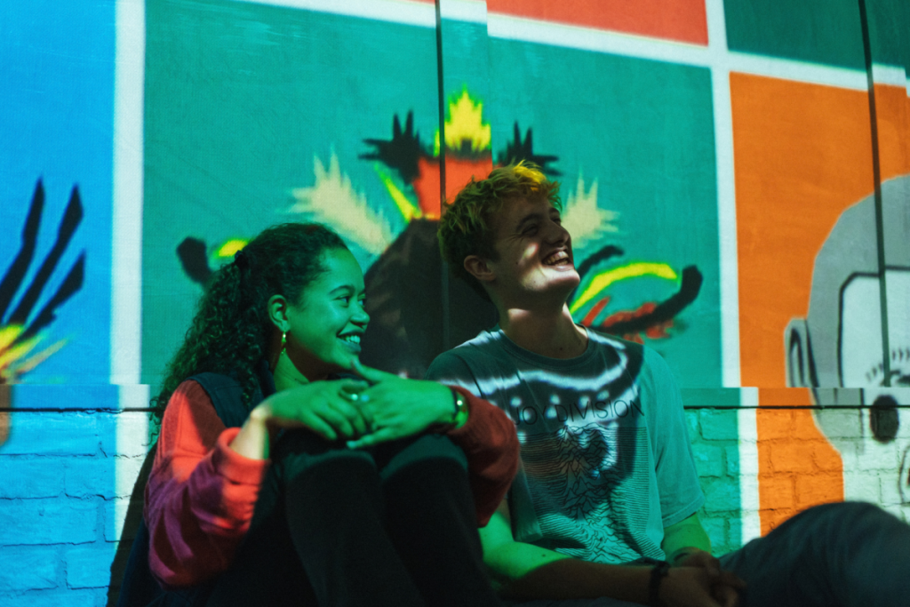 Deux jeunes visiteurs assis en souriant devant une fresque colorée à la Fabrique des Lumières.