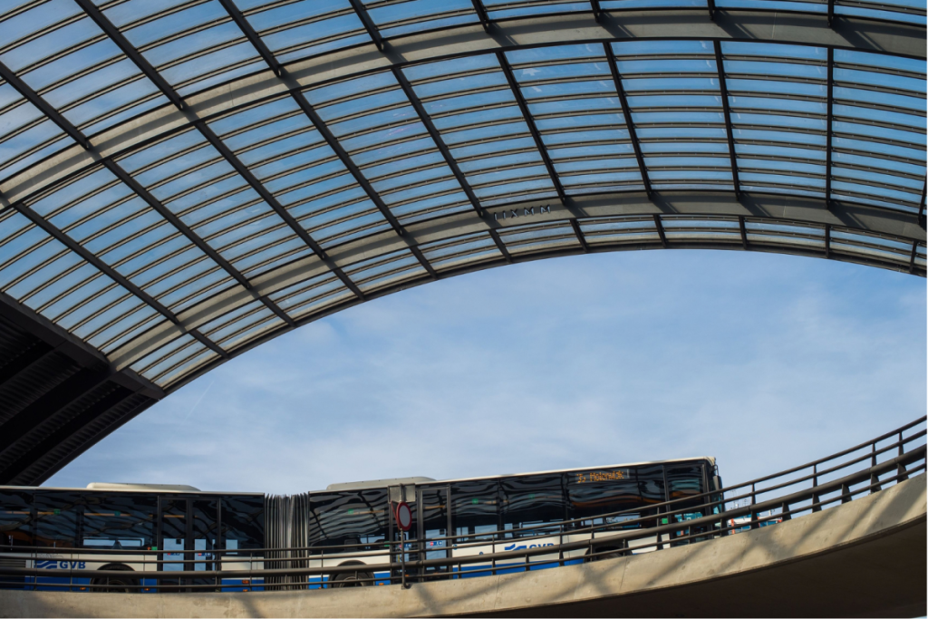 Ein blau-weißer GVB-Bus fährt über den Busbahnhof unter dem Glasdach des Amsterdamer Hauptbahnhofs.