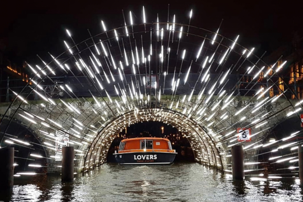 Ein Rundfahrtboot von „Lovers“ fährt während des Amsterdam Light Festivals unter einer beeindruckend beleuchteten Brücke hindurch.
