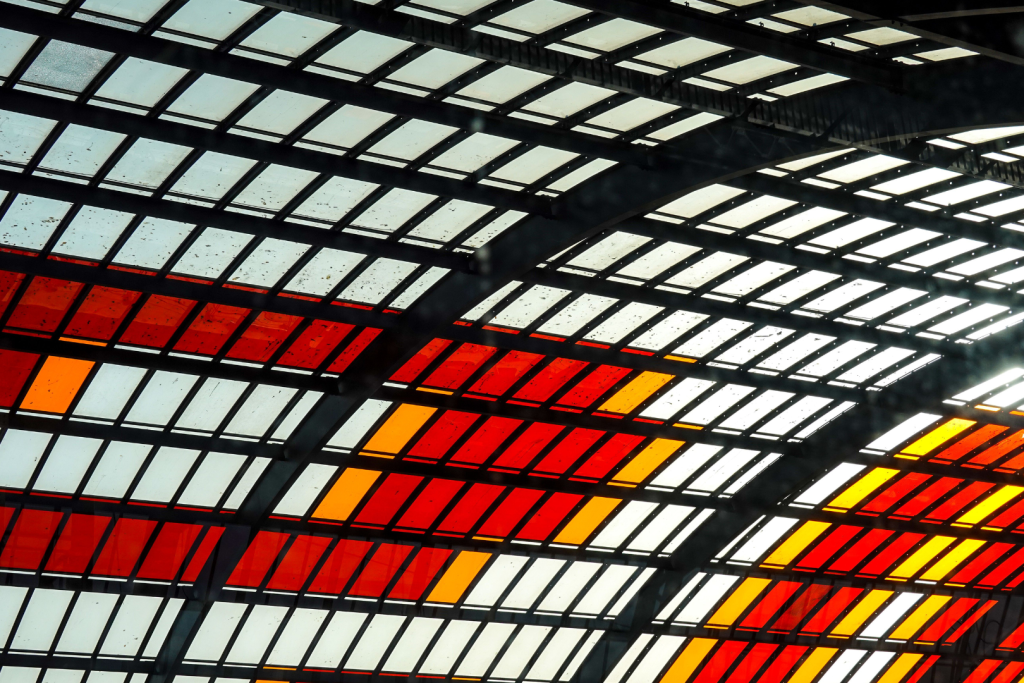 Abstraktes Foto der modernen Glasüberdachung der IJ-Halle am Amsterdamer Hauptbahnhof aus der Untersicht, mit einem Muster aus roten, orangefarbenen und weißen Glasplatten in einem Stahlrahmen.