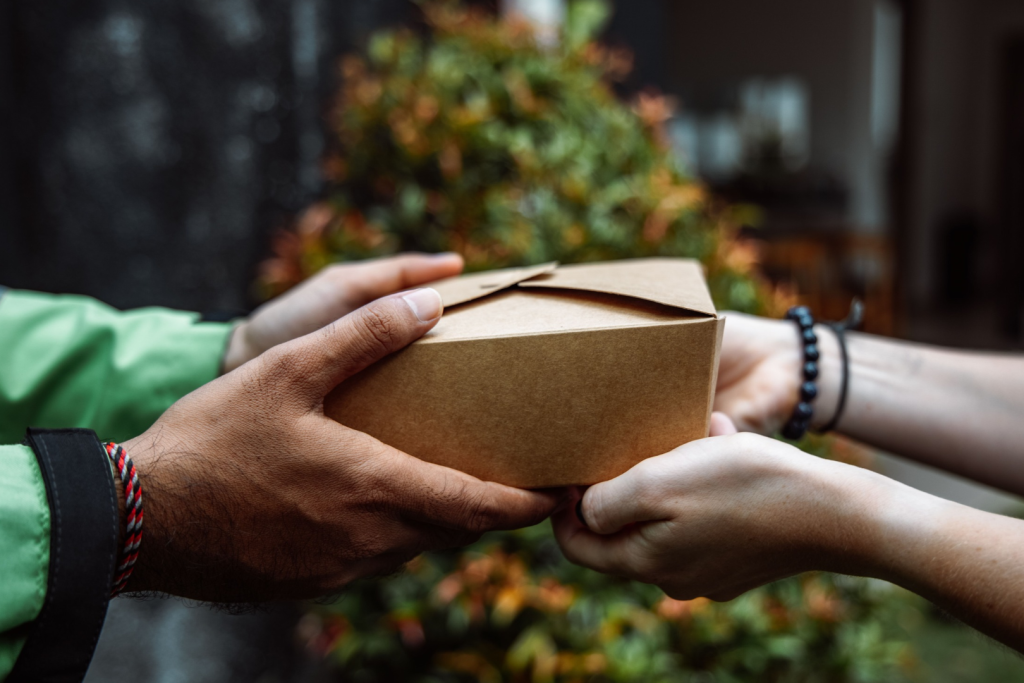 Zwei Hände reichen eine umweltfreundliche Mahlzeitbox aus Pappe an eine andere Person draußen weiter