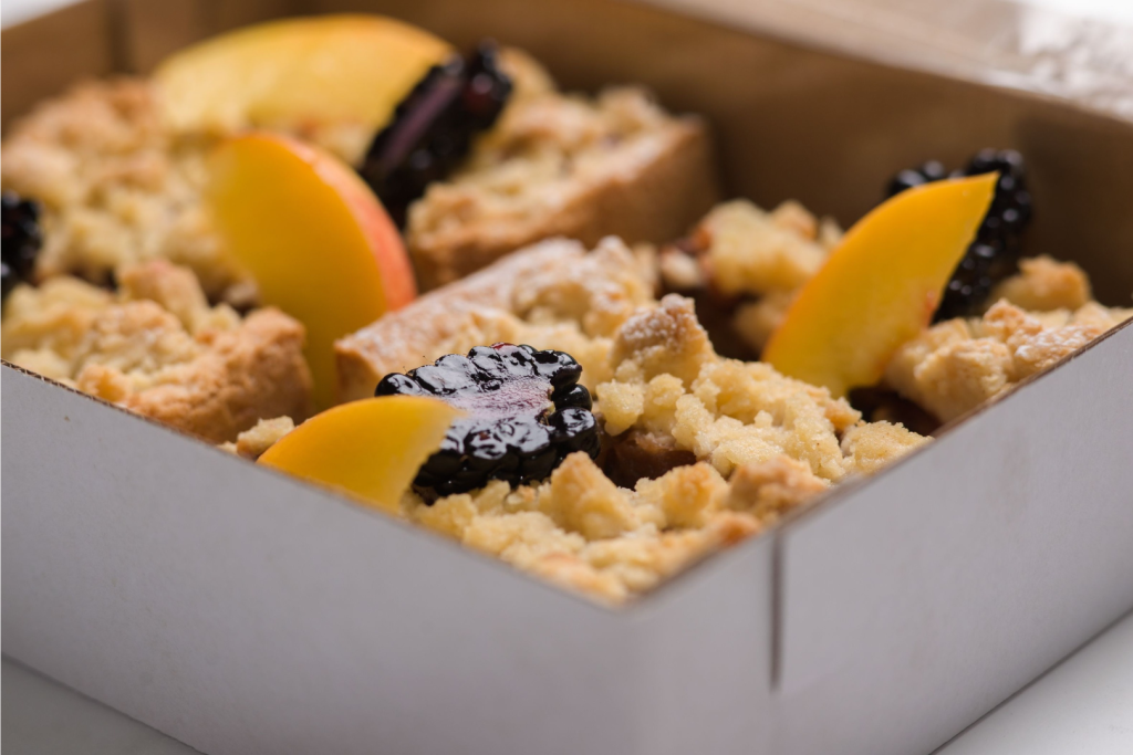 Frischgebackenes Blechgebäck mit Streuseln, frischen Pfirsichscheiben und Brombeeren in einer Pappverpackung