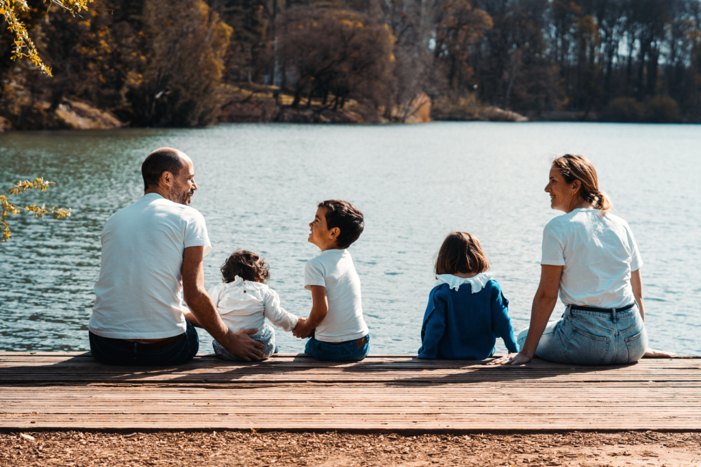 Eine Familie mit drei Kindern und Eltern, die auf einem Holzsteg am Wasser sitzen
