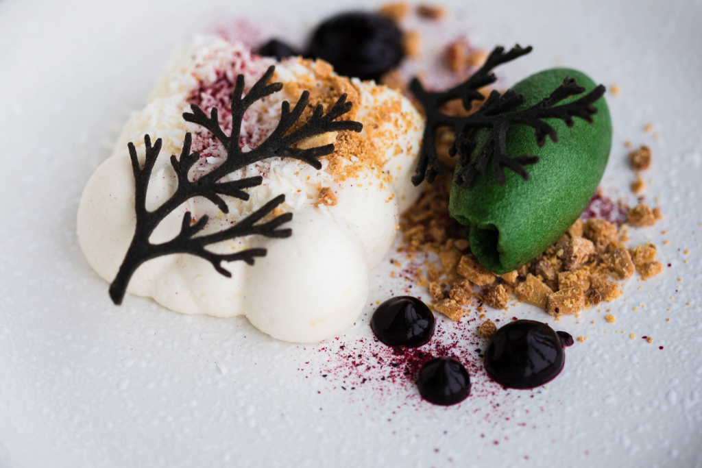 Dessert mit weißem Schaum, grünem Sorbet, knusprigem Crumble und Schokoladendekor.