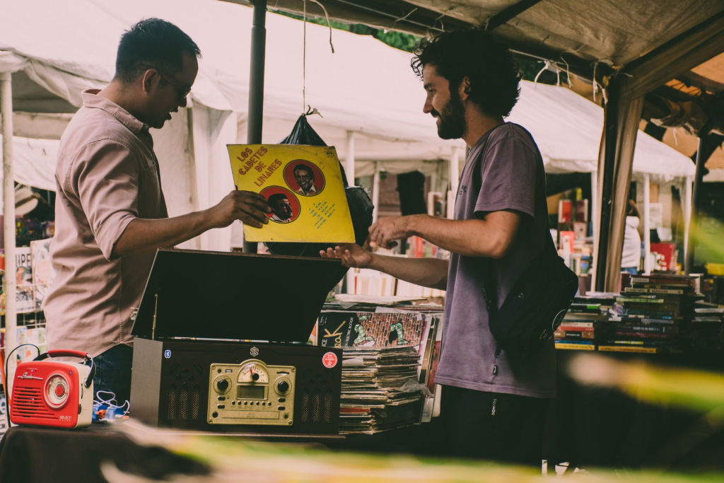 Zwei Männer betrachten ein gelbes LP-Cover an einem Stand auf einem überdachten Vinylmarkt mit Vintage-Plattenspielern.