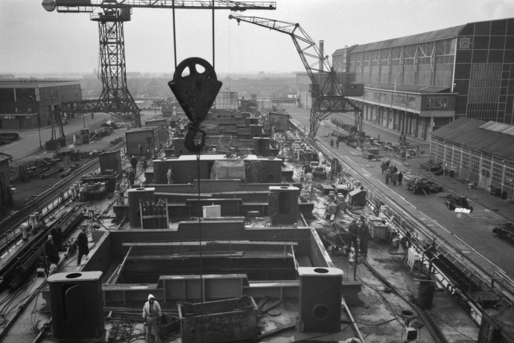 Historisches Schwarz-Weiß-Foto der aktiven NDSM-Werft mit Krananlagen, Arbeitern und im Bau befindlichen Schiffsrümpfen.