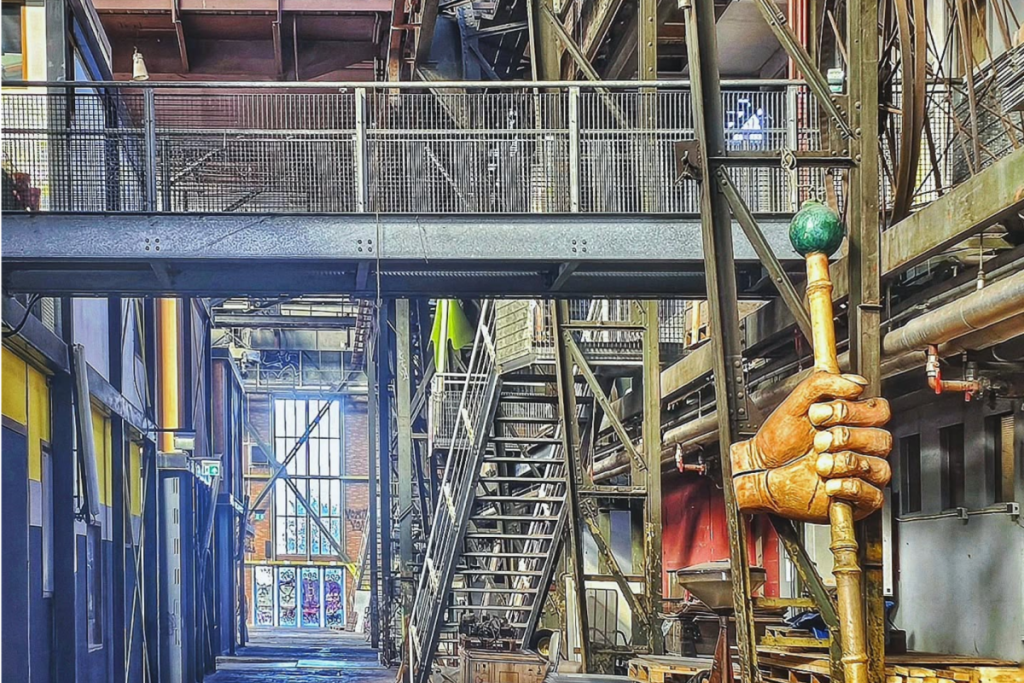 Das industrielle Stahlinterieur der NDSM-Halle mit Treppen, einer Laufbrücke und einer auffälligen hölzernen Kunstskulptur in Form einer Hand.