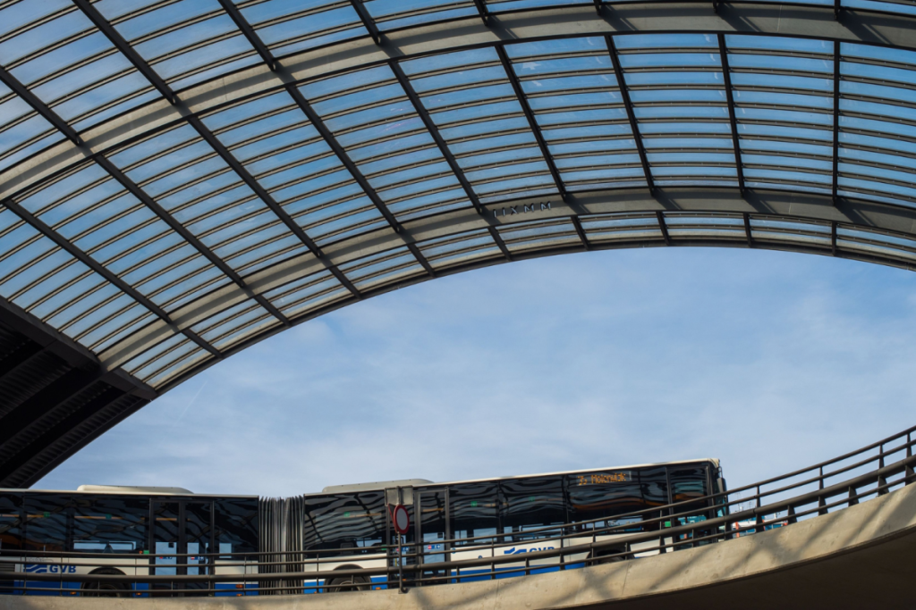 Ein blau-weißer GVB-Stadtbus fährt auf einer erhöhten Busspur unter dem Vordach eines Bahnhofs.