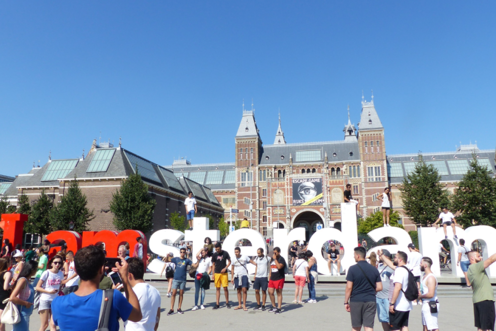 Besucher auf dem sonnigen Museumplein mit dem Rijksmuseum und den „I amsterdam“-Buchstaben im Hintergrund