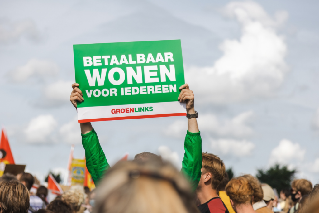 Ein Demonstrant hält während des Wohnprotests ein grünes Schild mit der Aufschrift „Bezahlbares Wohnen für alle“ hoch.