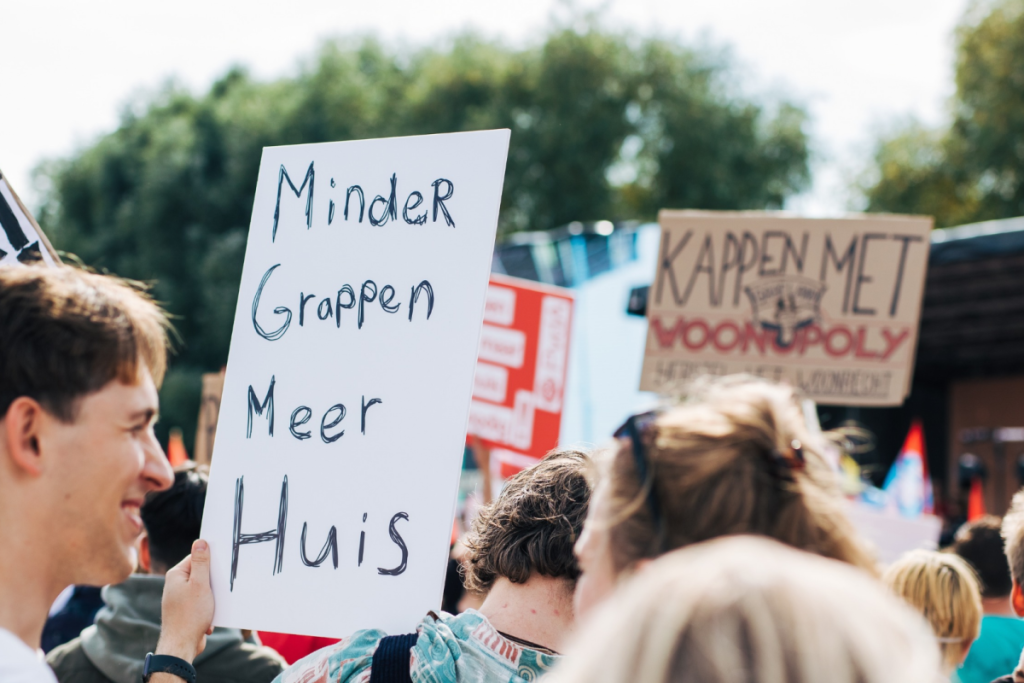 Ein Demonstrant hält während einer Wohnungsdemonstration ein handgeschriebenes Schild mit der Aufschrift „Weniger Witze, mehr Wohnraum“ hoch. JPG