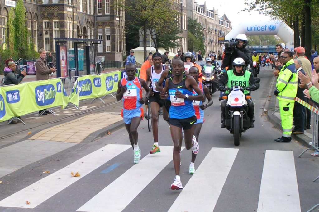 Die Spitzengruppe des Amsterdamer Marathons läuft über einen Zebrastreifen, begleitet von Motorrädern der Organisation und Zuschauern hinter den Absperrgittern.