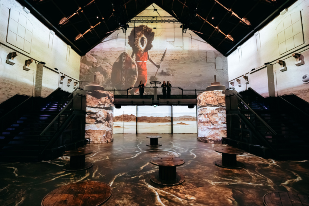Wandprojektion eines Kriegers mit Schild und Speer in einer Wüstenlandschaft während der Ausstellung von Jimmy Nelson in der Fabrique des Lumières.