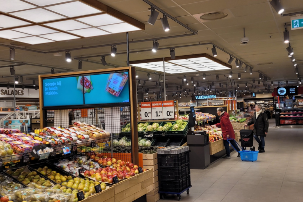 Blick in einen großen Albert-Heijn-Supermarkt mit gut gefüllten Regalen voller Obst und Gemüse, Bonus-Angebotsschildern und einkaufenden Kunden.