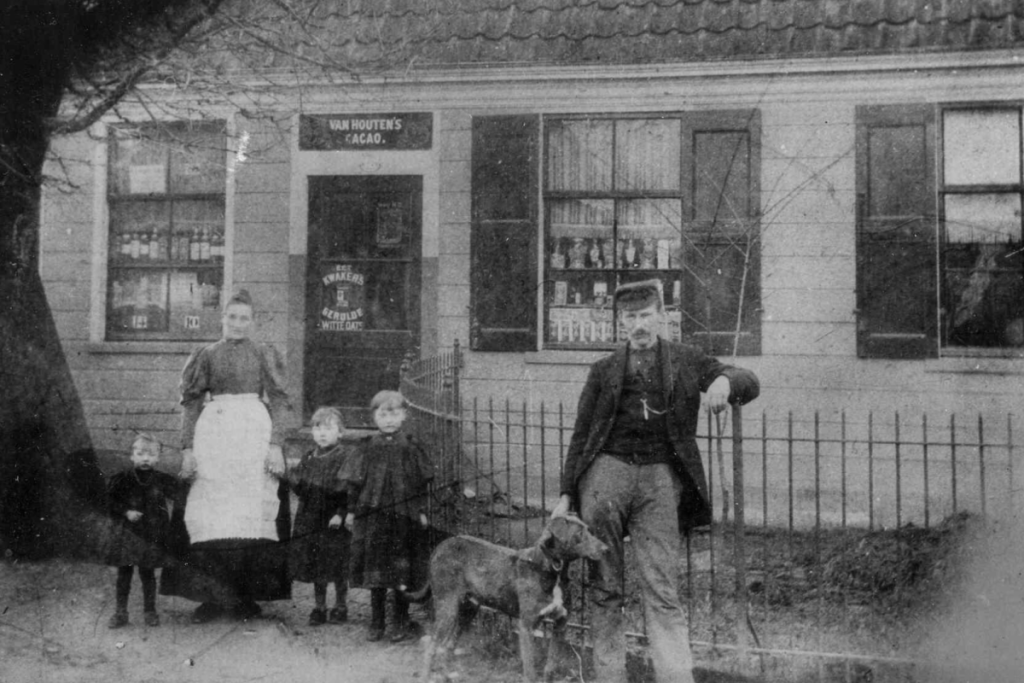 Historisches Schwarz-Weiß-Foto der Familie Heijn vor dem ursprünglichen kleinen Lebensmittelladen in Oostzaan mit einem Schild von „Van Houten’s Cacao“.