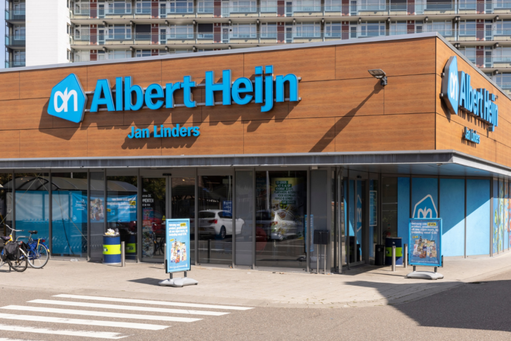 Außenansicht eines modernen Albert-Heijn-Supermarkts mit dem blauen Logo an einer Holzfassade und Glasschiebetüren.