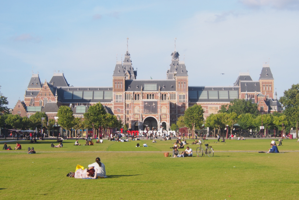 Ein sonniger Tag auf dem Museumplein in Amsterdam mit Besuchern auf der Wiese vor dem monumentalen Rijksmuseum.
