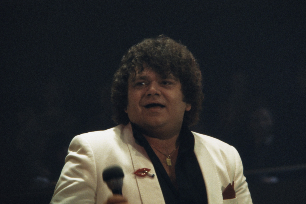 Sänger André Hazes tritt in einem weißen Sakko mit einem Mikrofon in der Hand auf der Bühne auf.