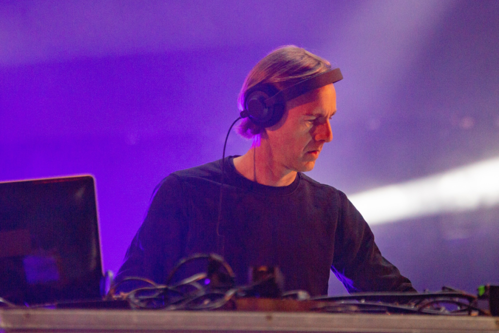 Techno-Legende Richie Hawtin steht mit Kopfhörern hinter seinem Mischpult und legt auf, während die Bühne in lila Licht getaucht ist.