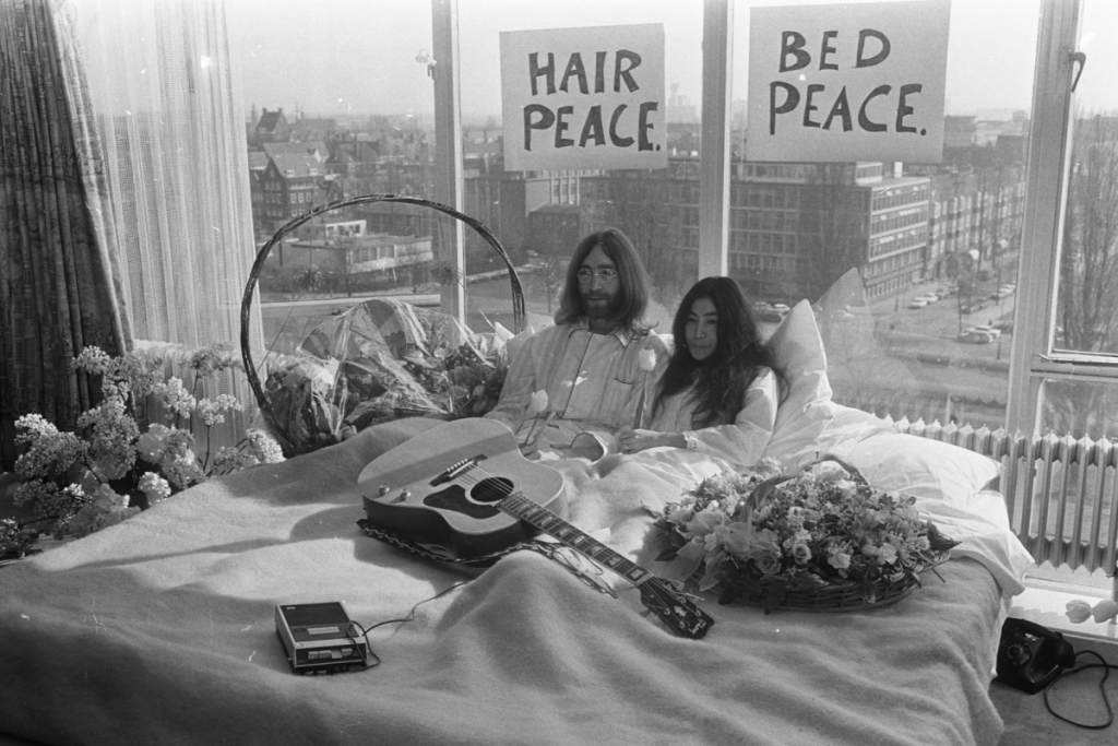 John Lennon und Yoko Ono im Bett während ihres „Bed-In“ für den Frieden im Hilton Hotel Amsterdam im Jahr 1969, mit Gitarre und den Postern „Bed Peace“ und „Hair Peace“.