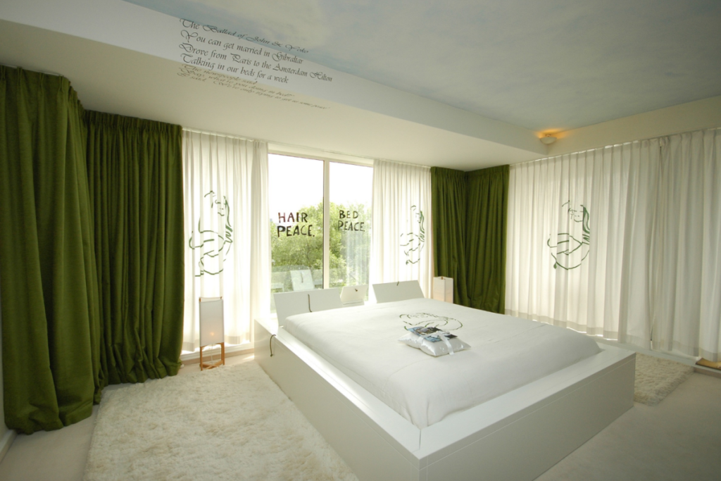 Innenansicht der renovierten John & Yoko-Suite im Hilton Amsterdam mit einem weißen Doppelbett, grünen Vorhängen und den ikonischen Schriftzügen „Bed Peace“ und „Hair Peace“ an den Fenstern.