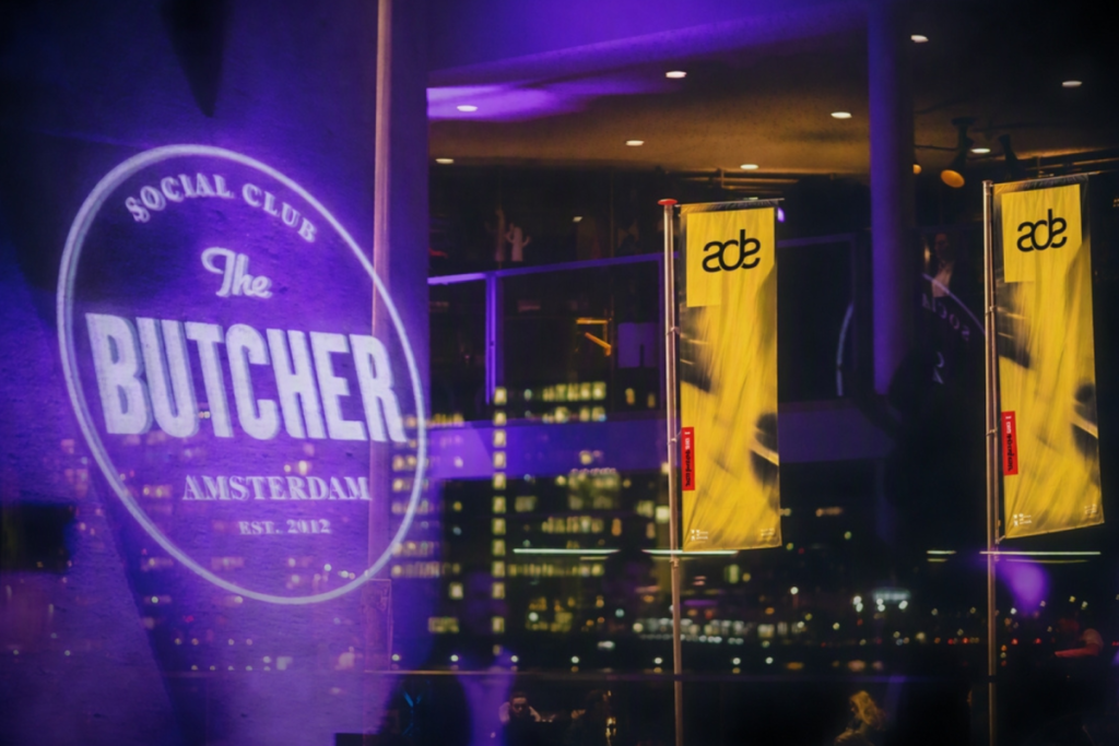 Das beleuchtete Logo des „The Butcher Social Club“ in Amsterdam, projiziert während des Amsterdam Dance Event auf ein Fenster, mit ADE-Flaggen im Hintergrund.