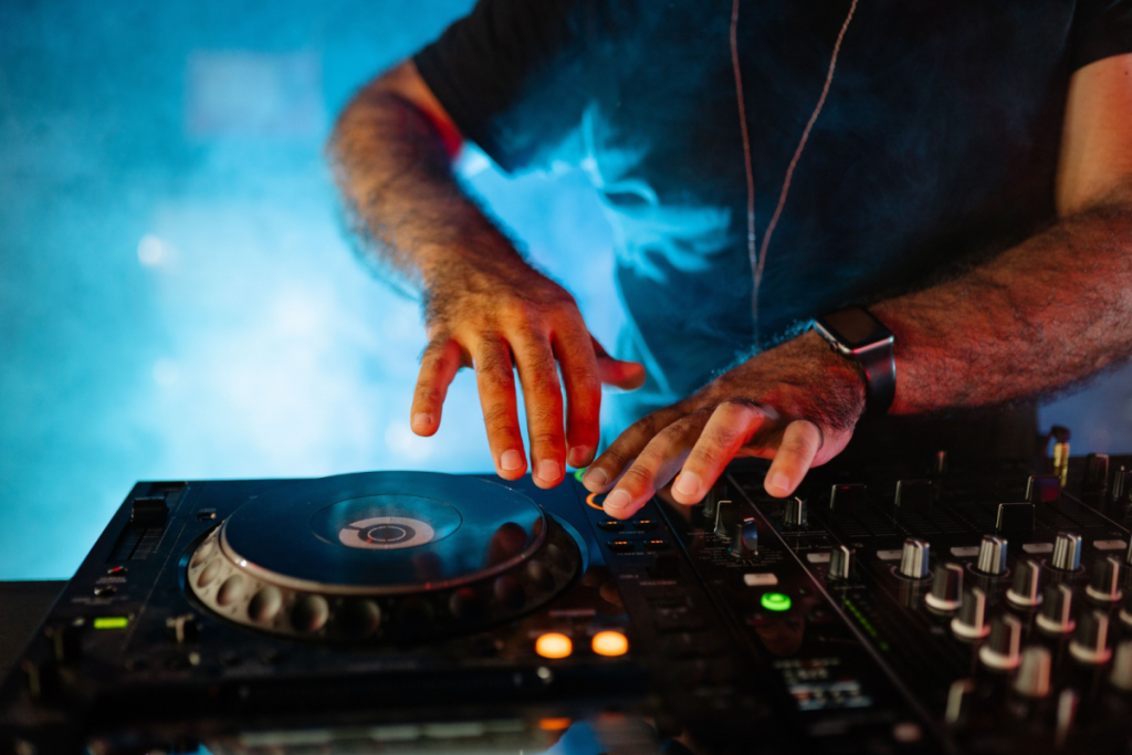 Nahaufnahme der Hände eines DJs, der während eines Live-Auftritts in einem stimmungsvoll beleuchteten Club ein Mischpult und einen Plattenspieler bedient.