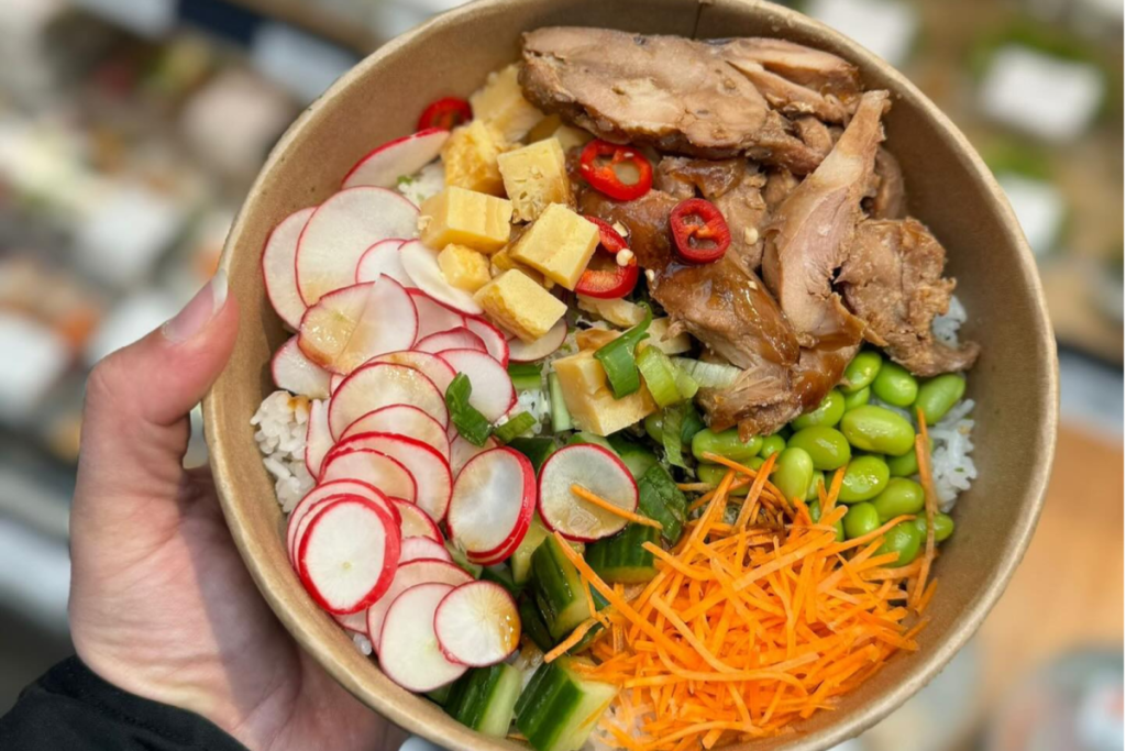 Frisch zubereitete Poké-Bowl vom Jumbo Foodmarkt mit Hähnchen, Radieschen, Edamame und Möhren in einer Pappschale.