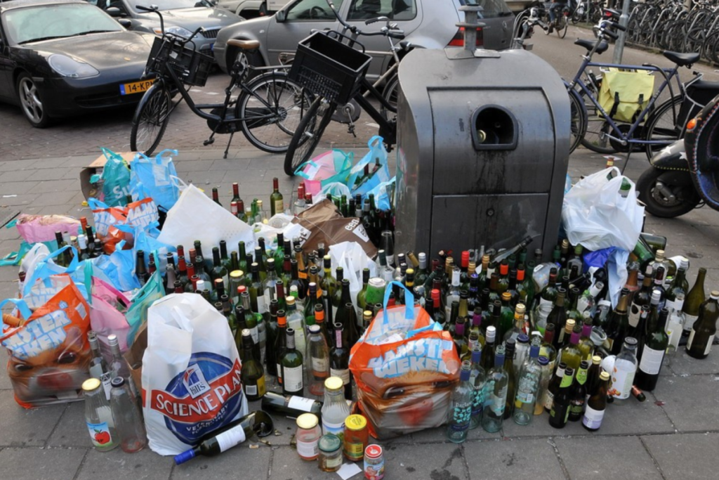 Contenitore per il vetro stracolmo per strada ad Amsterdam, circondato da bottiglie di vetro ammucchiate e sacchetti di plastica pieni di rifiuti abbandonati.