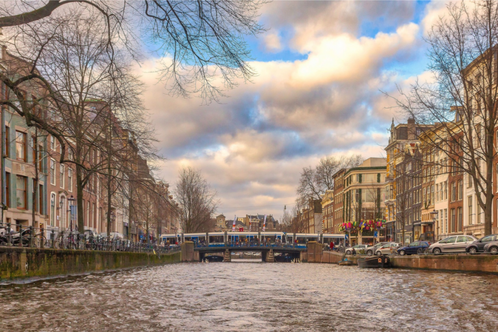 A blue-and-white tram crosses a stone bridge over an Amsterdam canal, surrounded by historic facades and bare trees in November.
