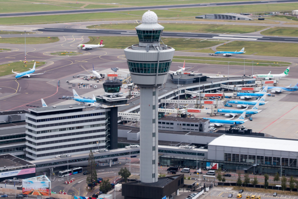 Luchtfoto van Schiphol Airport met de iconische verkeerstoren en vliegtuigen aan de gates.