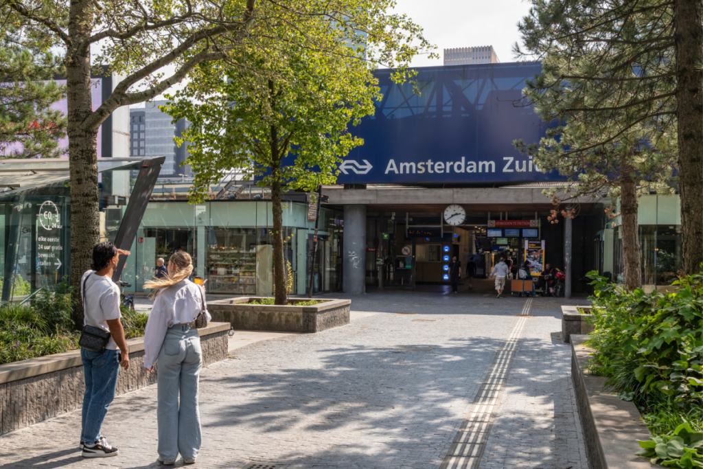 Außenansicht des Eingangs zum Bahnhof Amsterdam Zuid mit Reisenden auf dem Vorplatz, umgeben von Bäumen