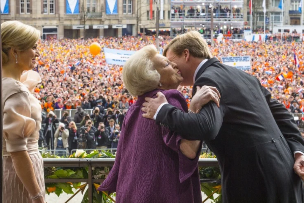 Koning Willem-Alexander kust Prinses Beatrix op het balkon van het Koninklijk Paleis Amsterdam tijdens de abdicatie in 2013, met Koningin Máxima en een volle Dam op de achtergrond