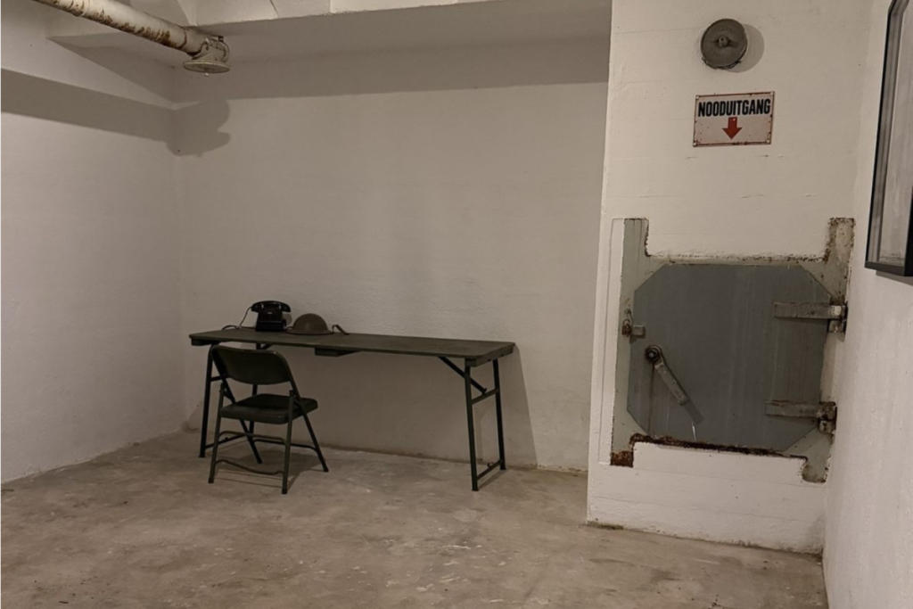 Ondergrondse ruimte van de atoombunker in Amsterdam met een stalen noodluik, witgeverfde betonmuren en een tafel met vintage telefoon en helm.