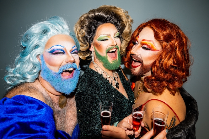 Three joyous drag queens, dressed in elaborate and elegant costumes with vibrant makeup, are sharing a light-hearted moment, laughing and toasting with glasses of wine