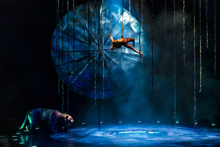 male acrobat hangs by one arm above a blue stage