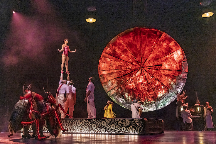 female acrobat stands on stilts, about to jump into a massive wheel