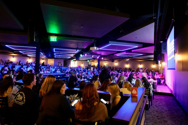a huge audience sitting at a comedy club with electric purple lights
