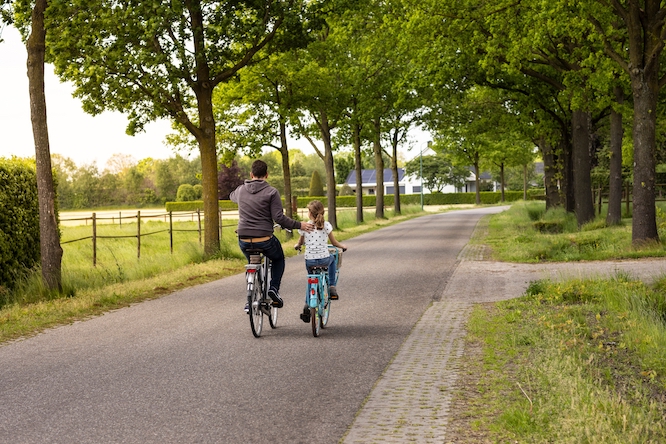Father and daughter cycling on a road in a rural area on the countryside, dad helping his girl with her bike and showing her the beautiful landscape and world. Parenthood, together, growing up concept