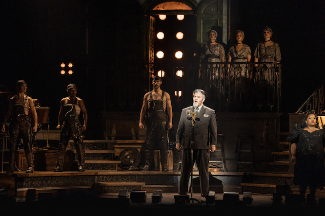 man in a suit standing on a dark stage surrounded by men dressed in workers outfits