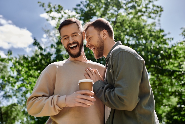 Two bearded men, a gay couple, share a laugh while enjoying a coffee together in a lush green park.